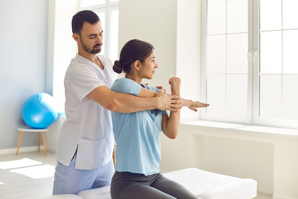 Male physiotherapist or chiropractor examining female patient's injured arm, stretching her muscles, helping her with medical exercise. Woman patient getting rehabilitation therapy in modern clinic